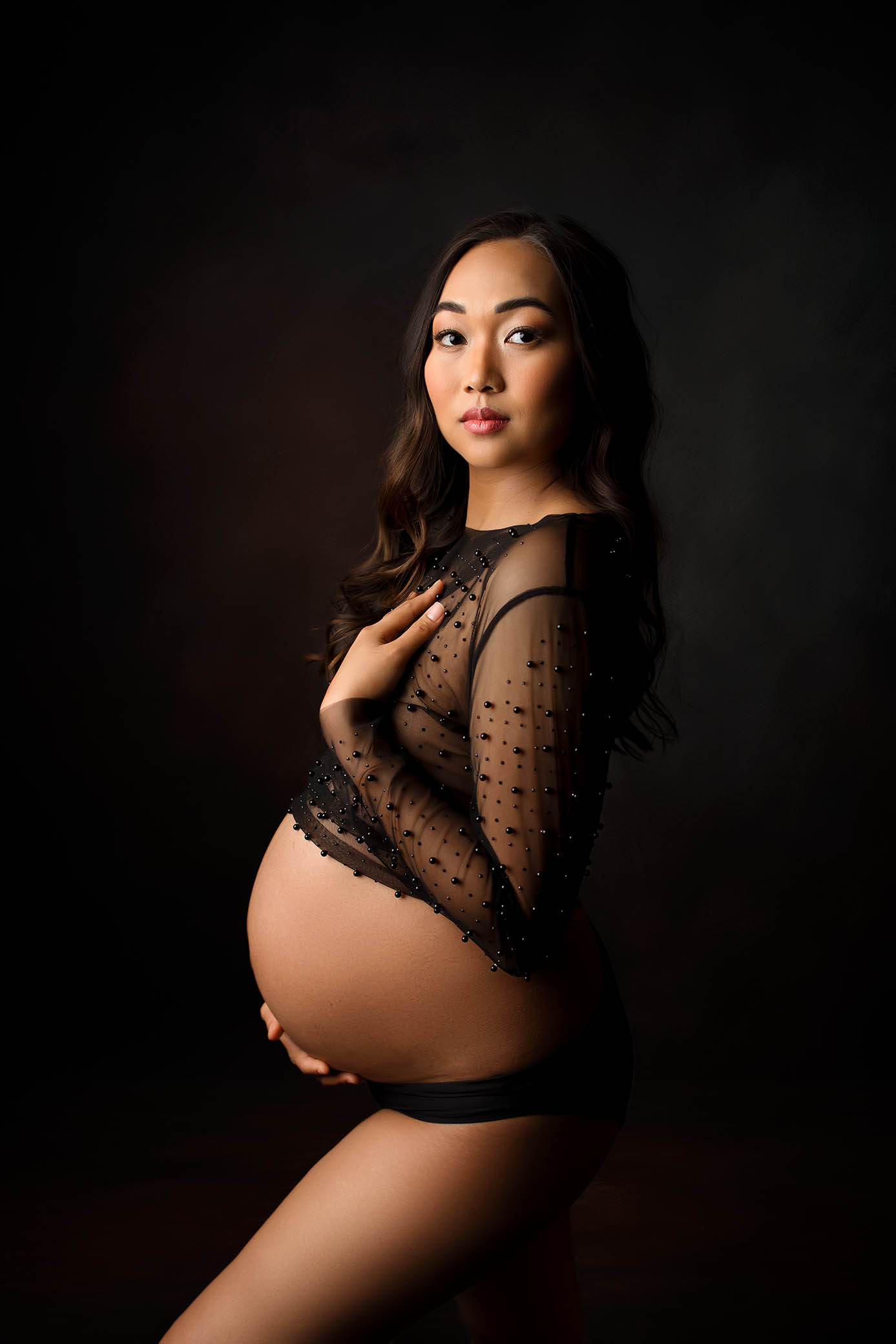 A pregnant woman with long dark hair poses in front of a dark background, wearing a sheer black top with decorative dots and black underwear, cradling her bare baby bump and looking confidently at the camera.