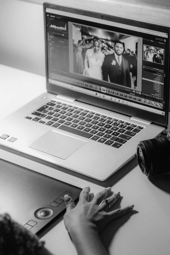 Person editing black-and-white wedding photo on laptop, with graphic tablet, stylus, and camera nearby.