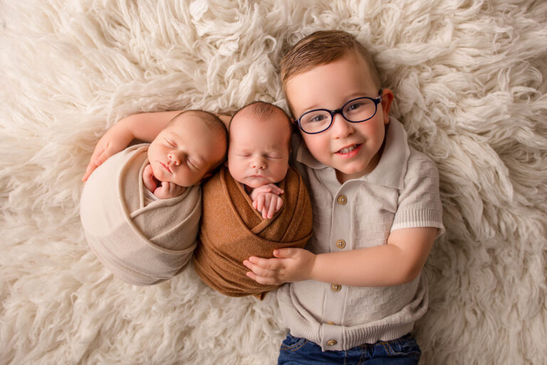 A young child with glasses smiles while lying on a fluffy white rug, gently hugging two swaddled newborn babies who are sleeping peacefully beside him.