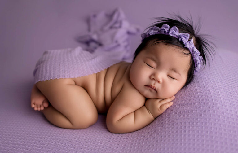 newborn photo session. A baby sleeps peacefully on a purple textured blanket, wearing a matching purple headband and draped with a lilac cloth, with a calm, relaxed expression.
