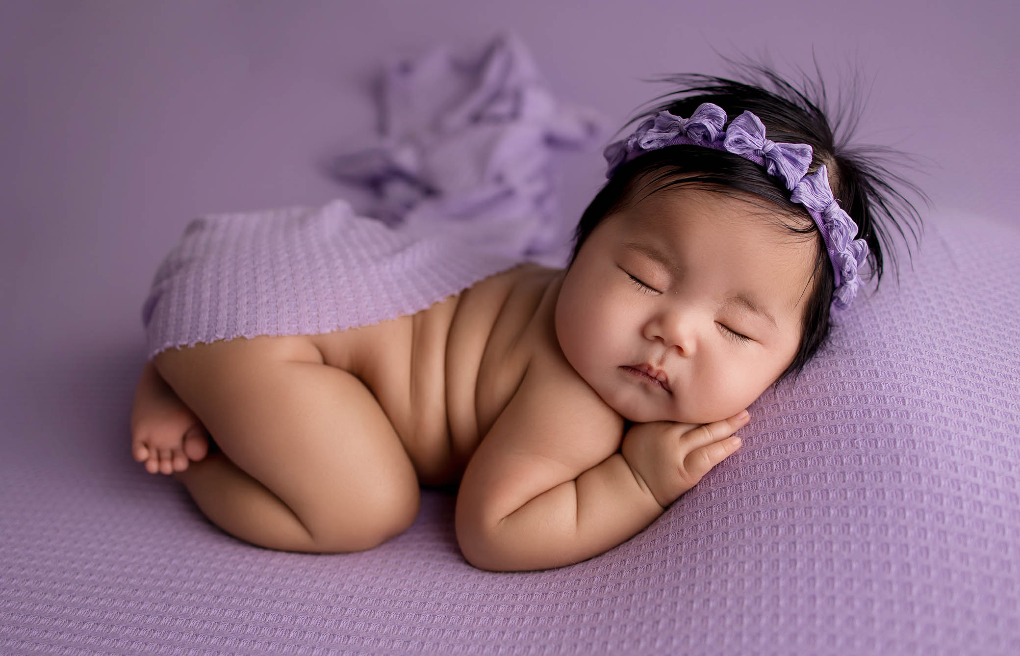 newborn photo session. A baby sleeps peacefully on a purple textured blanket, wearing a matching purple headband and draped with a lilac cloth, with a calm, relaxed expression.