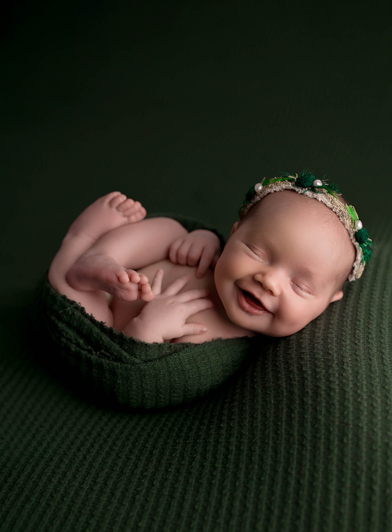 A smiling newborn baby with closed eyes lies on a dark green textured blanket, wrapped in a matching green swaddle. The baby wears a green and white floral headband.
