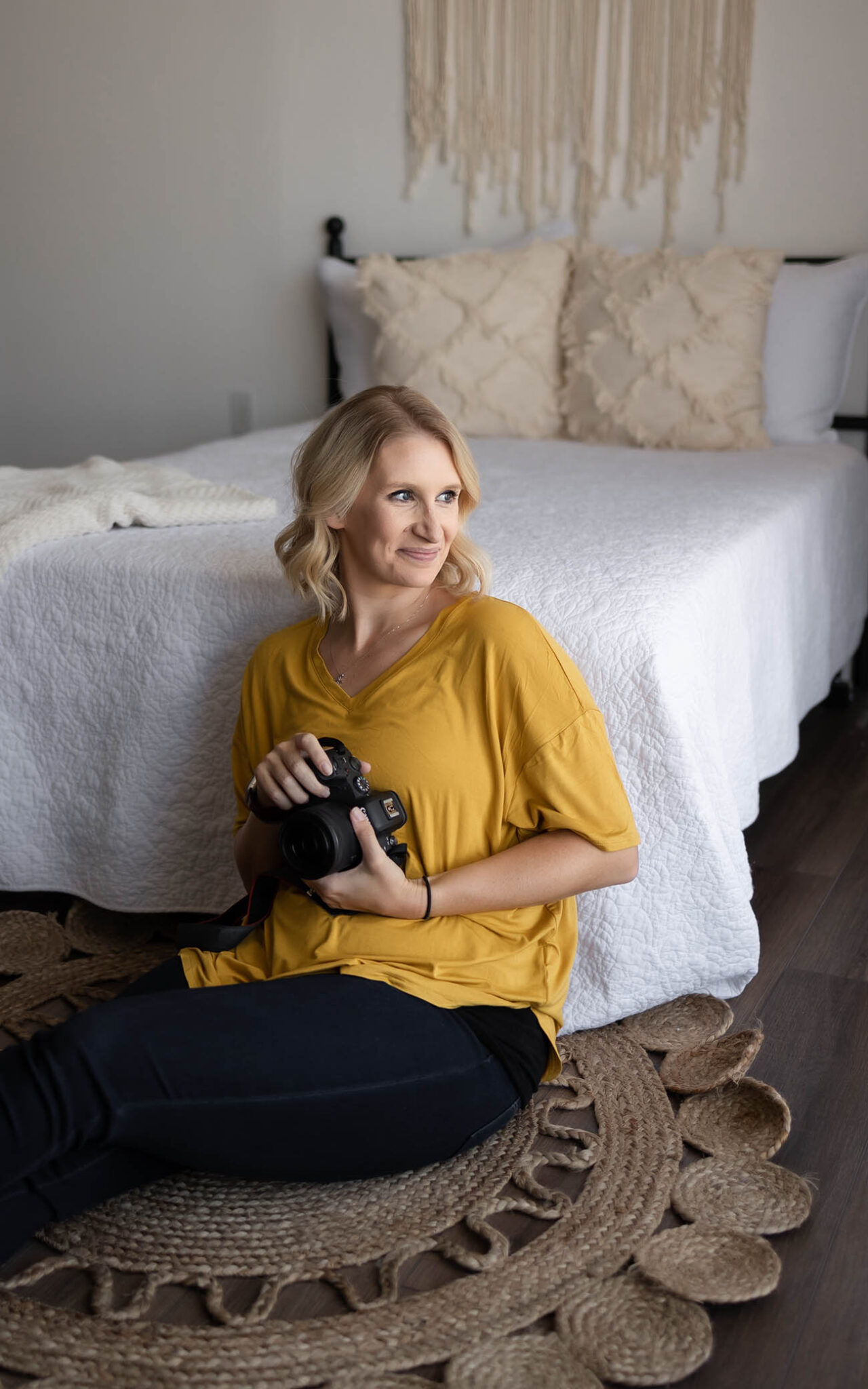 A woman with blonde hair wearing a yellow top sits on a textured rug, holding a camera, and smiling while looking to the side. Behind her is a neatly made bed—perfectly styled like a phoenix maternity photographer’s serene studio space.