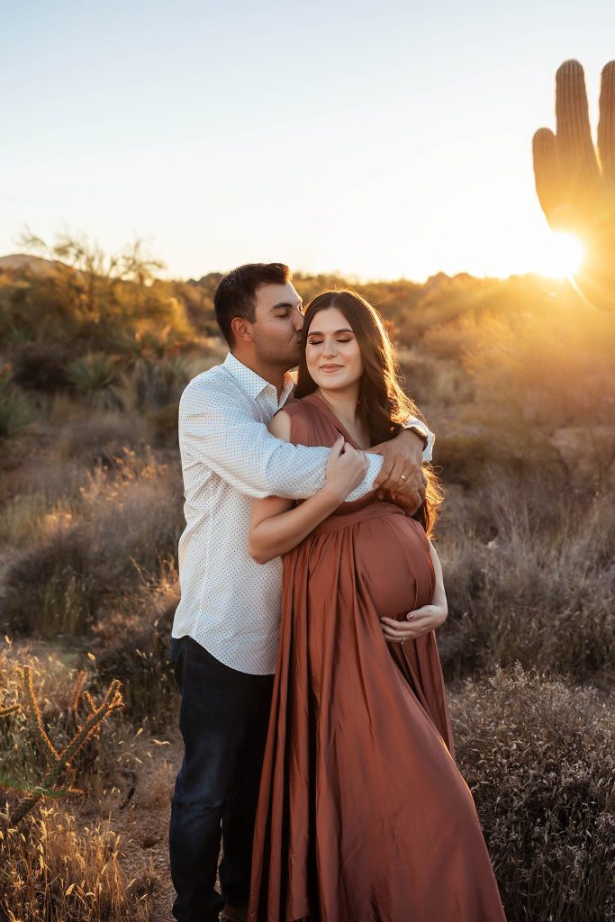 A man hugs and kisses a pregnant woman in a flowing rust dress at sunset in a desert landscape, with a cactus and sunlight in the background—captured by a talented family photographer Phoenix AZ.