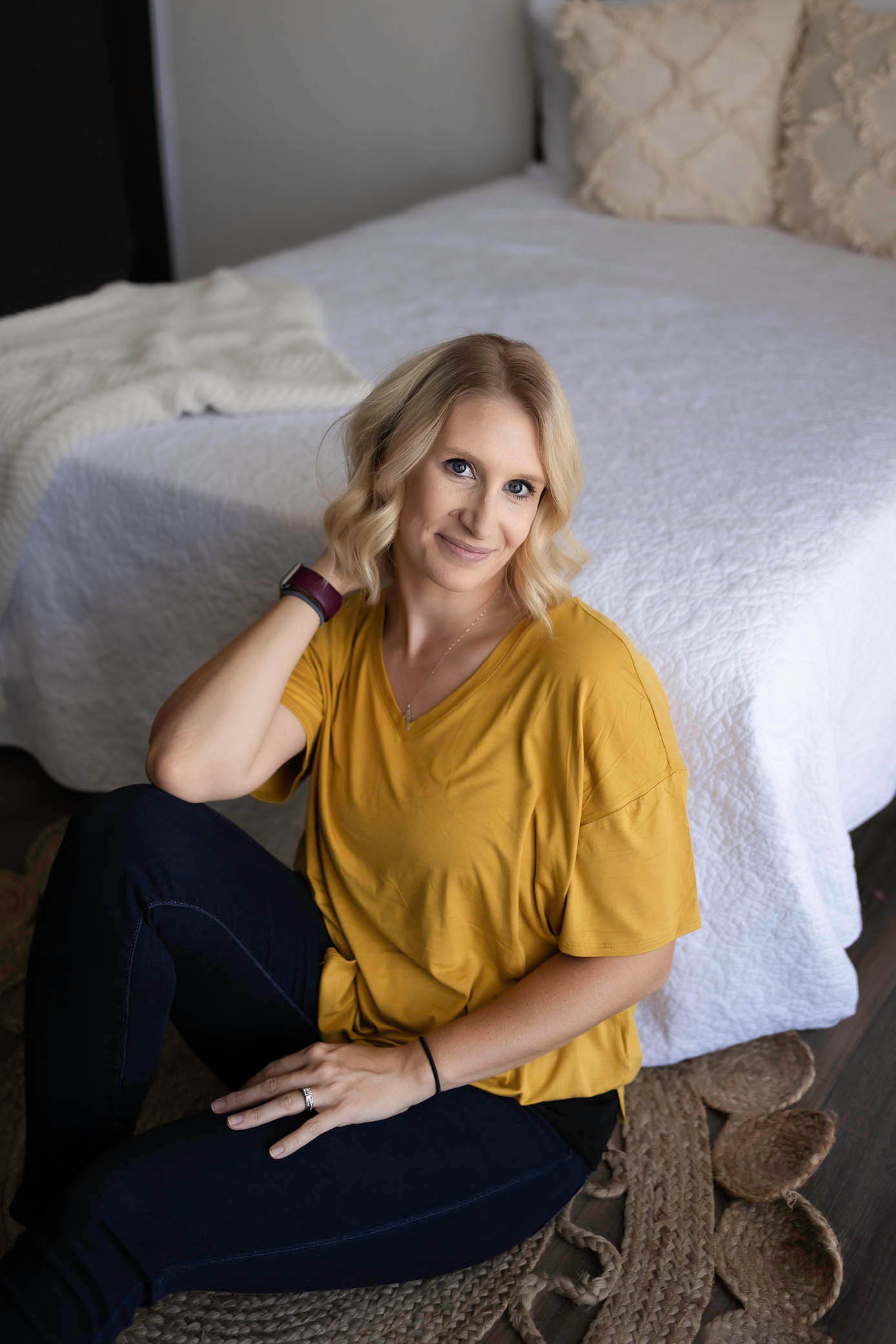 A woman with blonde hair in a yellow shirt and dark jeans sits on the floor, smiling next to a bed with a white quilt and cream pillows. Captured by a Glendale maternity photographer, she wears a watch and ring, hand in her hair.
