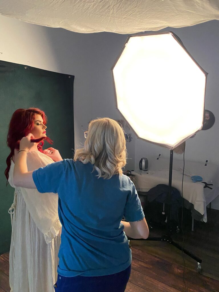 A woman in a white dress with red hair poses as another adjusts her look under a large studio light, showcasing the creative touch of a Scottsdale maternity photographer in a photography studio filled with unique props.