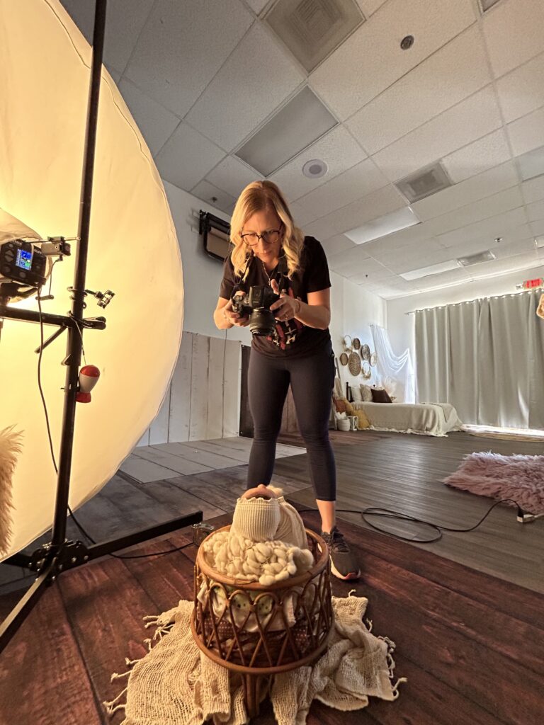 A family photographer Phoenix AZ leans forward with a camera, capturing precious photos of a swaddled baby in a basket in a well-lit studio with wooden floors, soft props, and a large lighting umbrella.