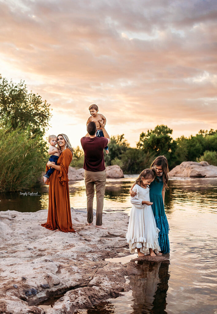 A family of five stands by a river at sunset, captured by a skilled phoenix newborn photographer. A woman in an orange dress holds a child, while a man in maroon lifts another. Two girls in blue and white dresses stand at the water's edge.