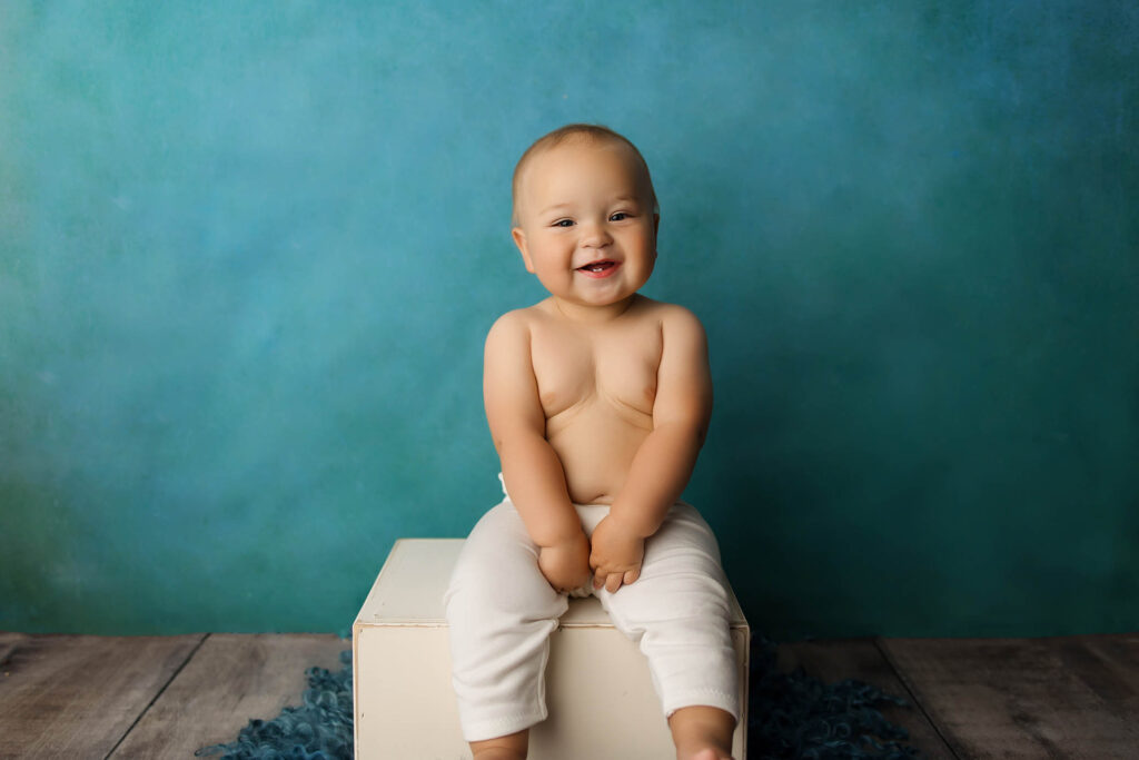 Smiling baby with light skin and short hair sits on a white box, wearing white pants, against a blue textured background on a wood floor—captured by a skilled Phoenix newborn photographer.