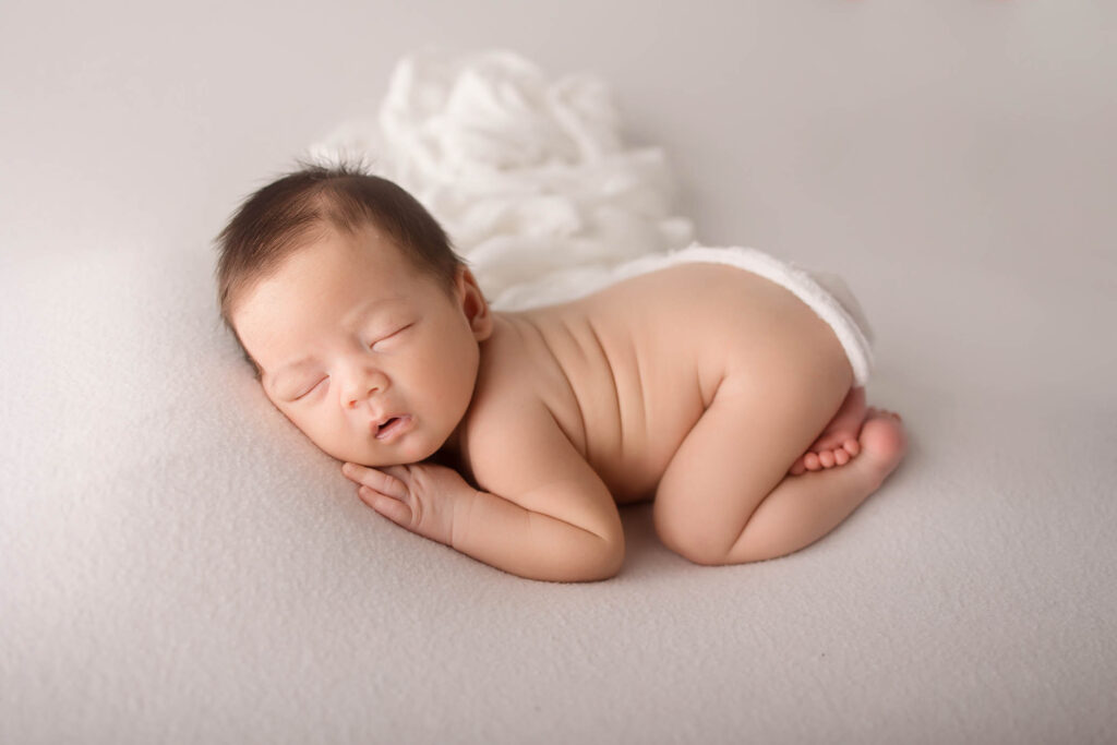 A newborn baby sleeps peacefully on a soft white surface, lying on their side with hands tucked under their cheek, wearing only a white diaper. Captured by a skilled Phoenix newborn photographer, a white blanket is softly draped in the background.