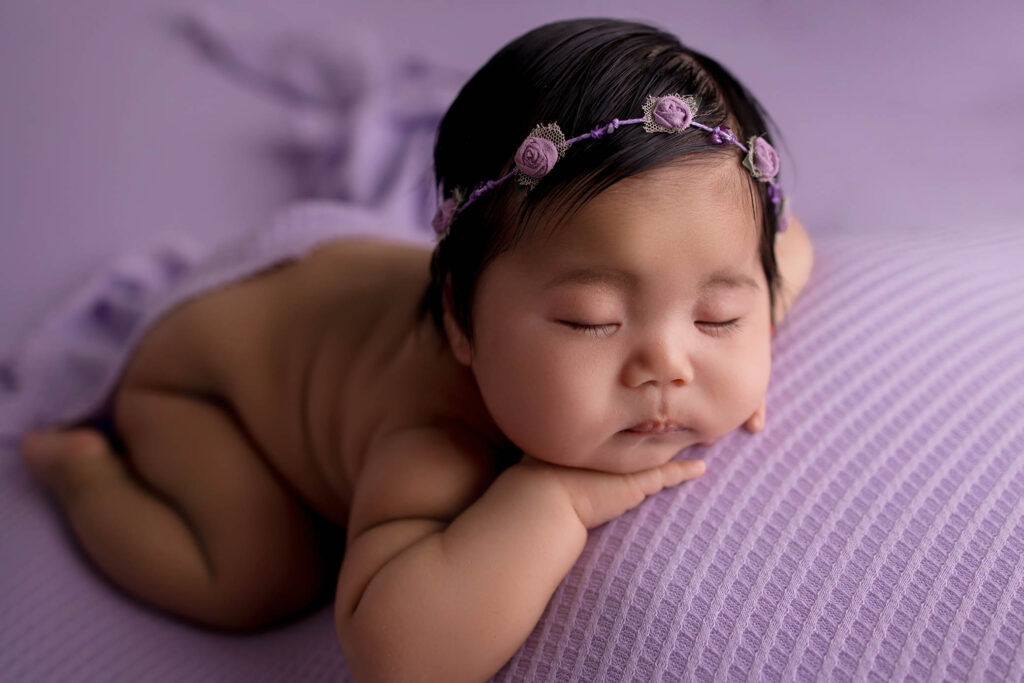 A baby with closed eyes sleeps peacefully on a textured purple blanket, wearing a lilac flower headband. Captured by a Phoenix newborn photographer, the baby rests their chin on their hands, surrounded by a soft, purple-toned background.