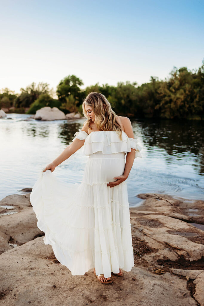 A pregnant woman in a white off-shoulder dress stands on rocks by a calm river, holding her belly with one hand and lifting her dress with the other, captured beautifully by a Phoenix maternity photographer amid trees and a blue sky.