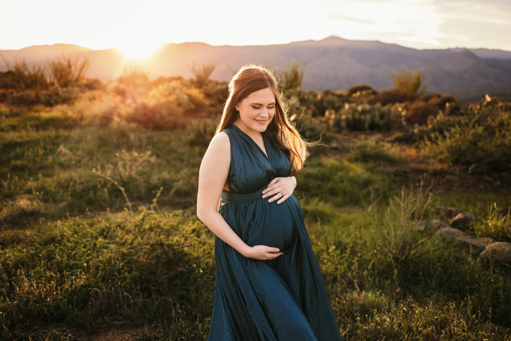 A pregnant woman in a flowing dark green dress stands in a grassy field at sunset, smiling and gently holding her belly, with mountains and golden sunlight in the background, captured beautifully by a Scottsdale maternity photographer.