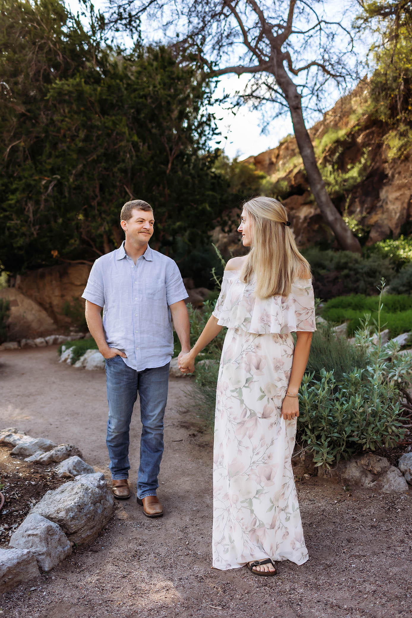 A couple walks hand in hand on a garden path, smiling at each other amid greenery and trees. The scene is beautifully captured by a family photographer Phoenix AZ, preserving this special moment in a natural setting.