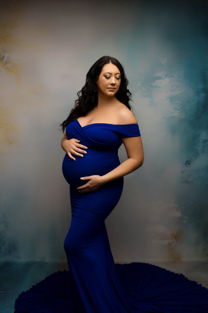 A pregnant woman with long dark hair wearing an off-the-shoulder, fitted royal blue gown cradles her baby bump and poses against a softly textured blue and cream background, beautifully captured by a Phoenix maternity photographer.