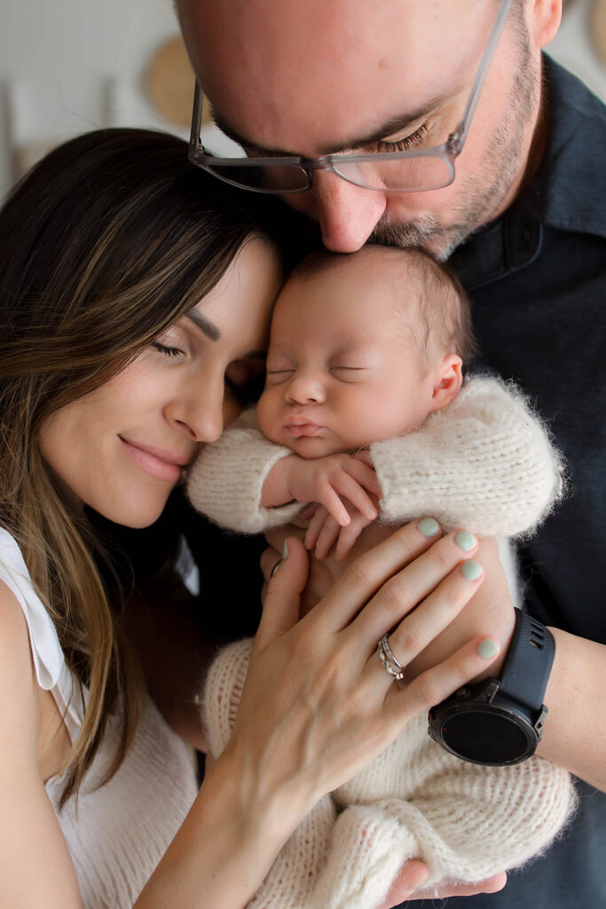 A woman and man gently hold and embrace a sleeping newborn baby, expressing love and tenderness. Captured by a skilled family photographer in Phoenix, AZ, the moment shows the woman resting her face on the baby as the man kisses the baby's head.