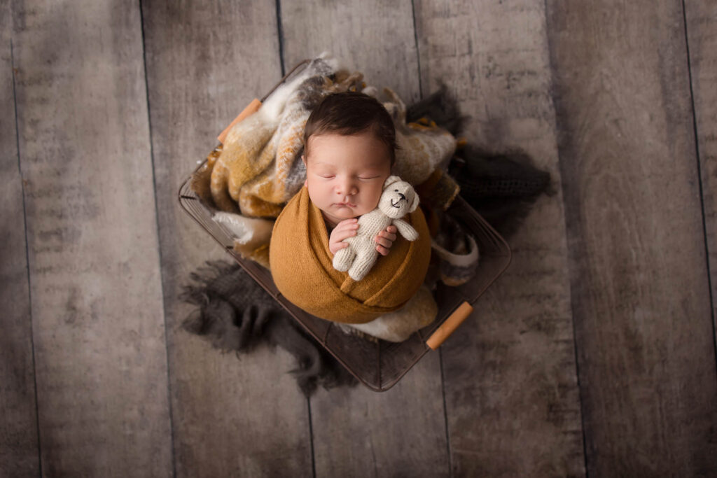 A newborn baby wrapped in a mustard yellow blanket lies in a basket on a wooden floor, peacefully sleeping with a small knitted teddy bear. Soft, neutral-toned blankets surround the little one—captured by a skilled Peoria maternity photographer.