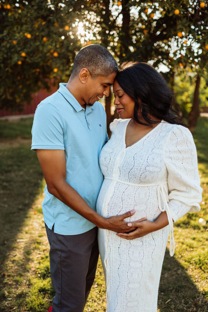 A smiling couple stands outdoors, touching foreheads affectionately. Captured by a Scottsdale maternity photographer, the woman cradles her pregnant belly as sunlight filters through trees behind them.