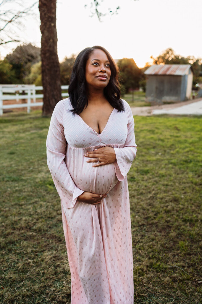A pregnant woman in a light pink, long-sleeved dress stands outside on grass, gently cradling her baby bump with both hands. Captured by a Peoria maternity photographer, she looks slightly upward with trees and a wooden structure in the background.