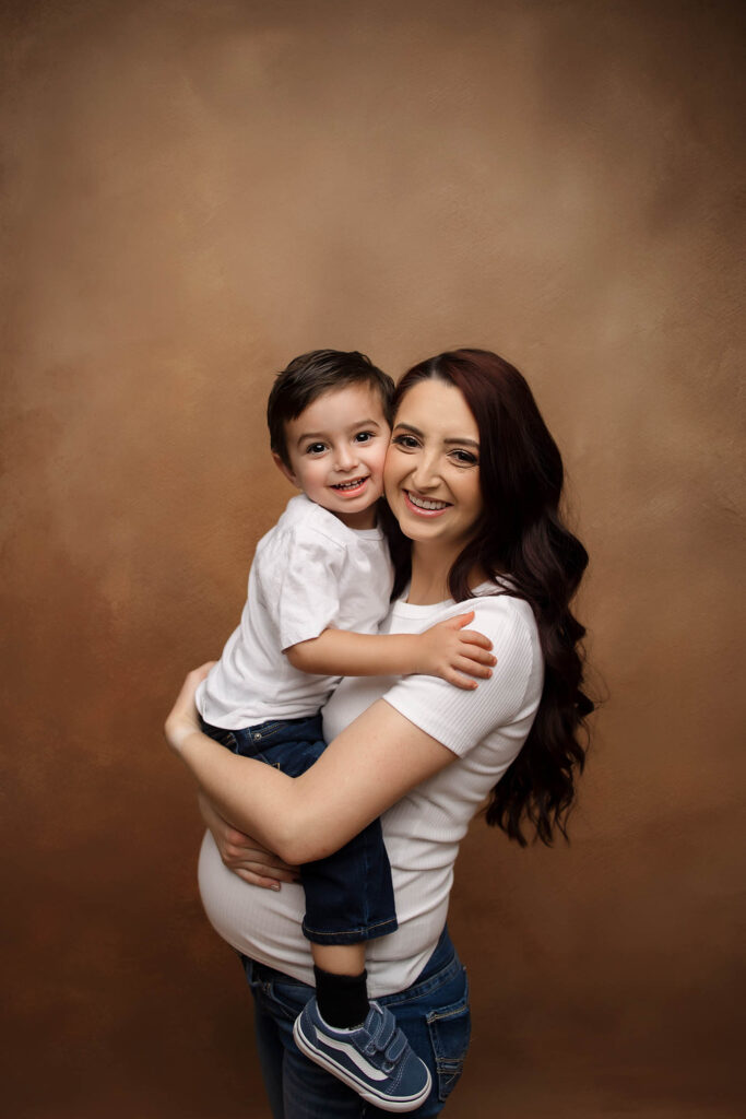 A smiling woman with long dark hair holds a happy young boy in her arms. Both wear white shirts and jeans, standing against a warm brown background. Captured by a Glendale maternity photographer, they look directly at the camera.