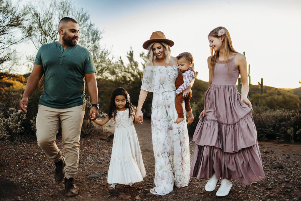 A family of five walks outdoors on a rocky path, surrounded by desert plants. Captured by a Scottsdale maternity photographer, the smiling adults and children enjoy a sunlit moment, with the woman in the center holding a toddler’s hand.