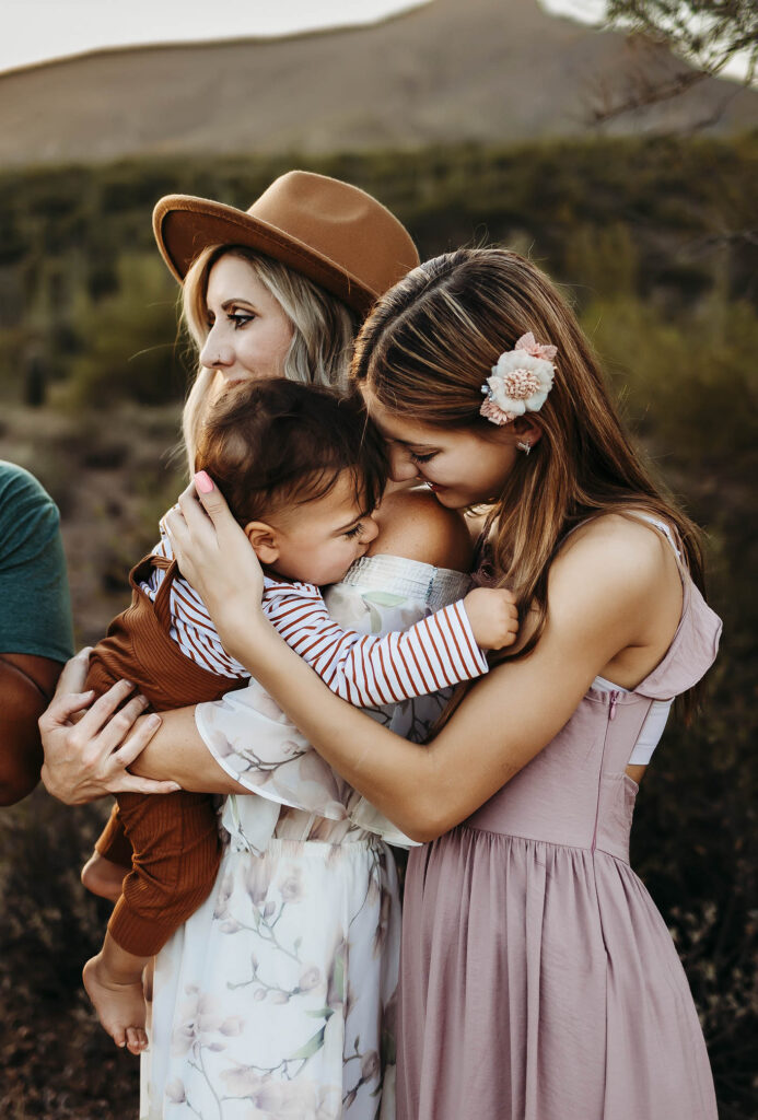 A woman in a floral dress and hat hugs a baby, while a girl in pink with a flower hair clip lovingly embraces the baby—all captured outdoors by a Phoenix maternity photographer, with mountains and lush greenery in the background.