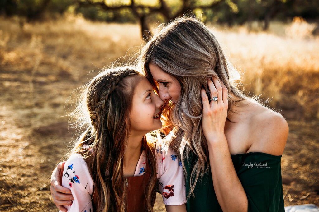A woman and a young girl smile lovingly at each other, touching foreheads while sitting outdoors in a sunlit, grassy field. Captured by a talented Phoenix maternity photographer, their happiness and connection shine through.