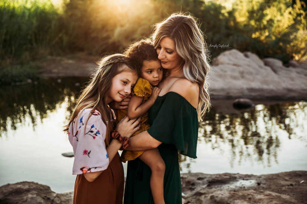 A woman lovingly hugs two young girls by a peaceful riverside at sunset, with warm sunlight illuminating their faces and tall greenery in the background—captured beautifully by a Phoenix maternity photographer.