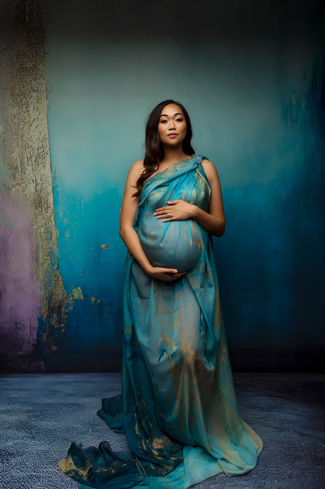 A pregnant woman in a flowing blue and gold gown stands against a textured blue background, cradling her belly confidently—captured perfectly by a Scottsdale maternity photographer.