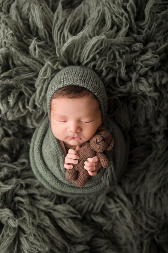 A newborn baby wrapped in a green knit blanket and bonnet sleeps peacefully on a fluffy dark green rug, holding a small brown knitted teddy bear. Captured with care by a Phoenix newborn photographer.