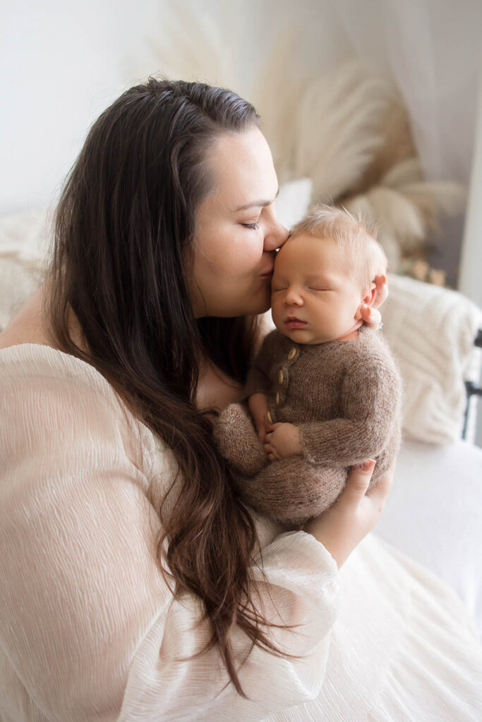A woman with long dark hair kisses the forehead of a sleeping newborn dressed in a cozy brown outfit as they cuddle in a softly lit, neutral-toned room—captured by a skilled Phoenix newborn photographer.