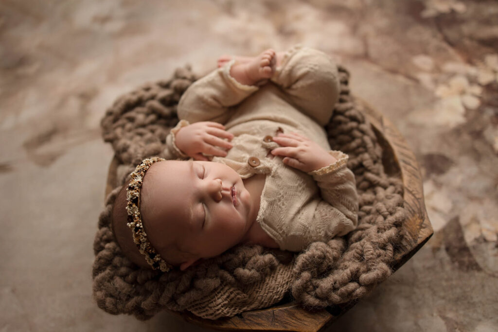 A newborn baby wearing a beige outfit and floral headband sleeps peacefully on a chunky brown knit blanket in a wooden bowl, beautifully captured by a Phoenix newborn photographer.
