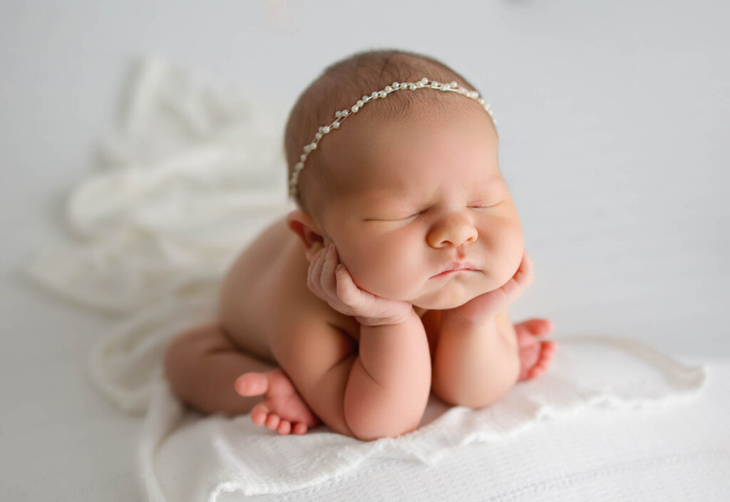 A newborn baby with a delicate beaded headband is posed with elbows on a soft white blanket, chin resting in hands, and eyes closed—captured in pure innocence and peace by a skilled Phoenix newborn photographer.