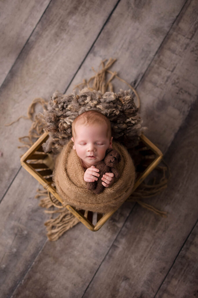 A newborn baby is swaddled in a brown wrap, lying in a wooden basket on a textured blanket, holding a small stuffed animal, peacefully sleeping—captured by a Peoria maternity photographer from above on a wooden floor.