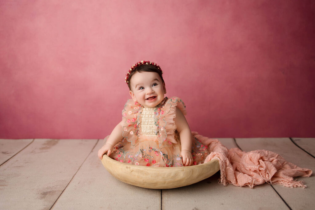 Smiling baby in a peach-colored, ruffled dress and floral headband sits in a wooden bowl on a wooden floor with a pink textured backdrop, beautifully captured by a Phoenix newborn photographer.