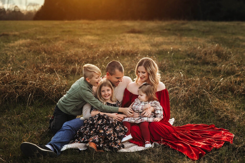 A family of five sits on a blanket in a grassy field at sunset, smiling and embracing each other. Captured by a Scottsdale maternity photographer, the mother glows in a long red dress as the father and children laugh together, radiating happiness.