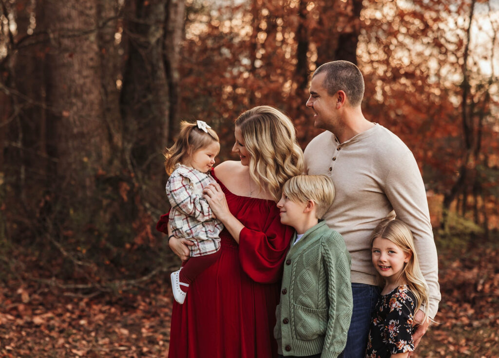 A family of five stands outdoors in autumn. Captured by a family photographer in Phoenix, AZ, the mother in a red dress holds a young girl, while the father and two older children—smiling at each other—complete this warm, joyful moment.