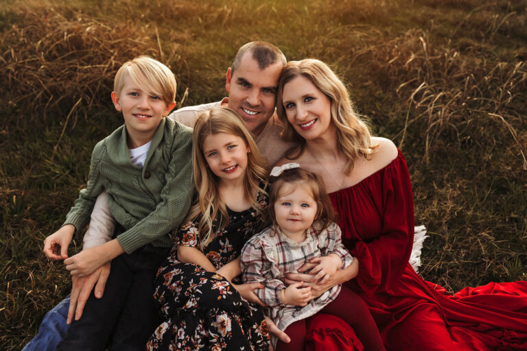 A family of five sits closely together on grass at sunset, smiling for the camera. Captured by a talented family photographer Phoenix AZ, their casual, earthy-toned outfits and warm expressions create a timeless outdoor portrait.