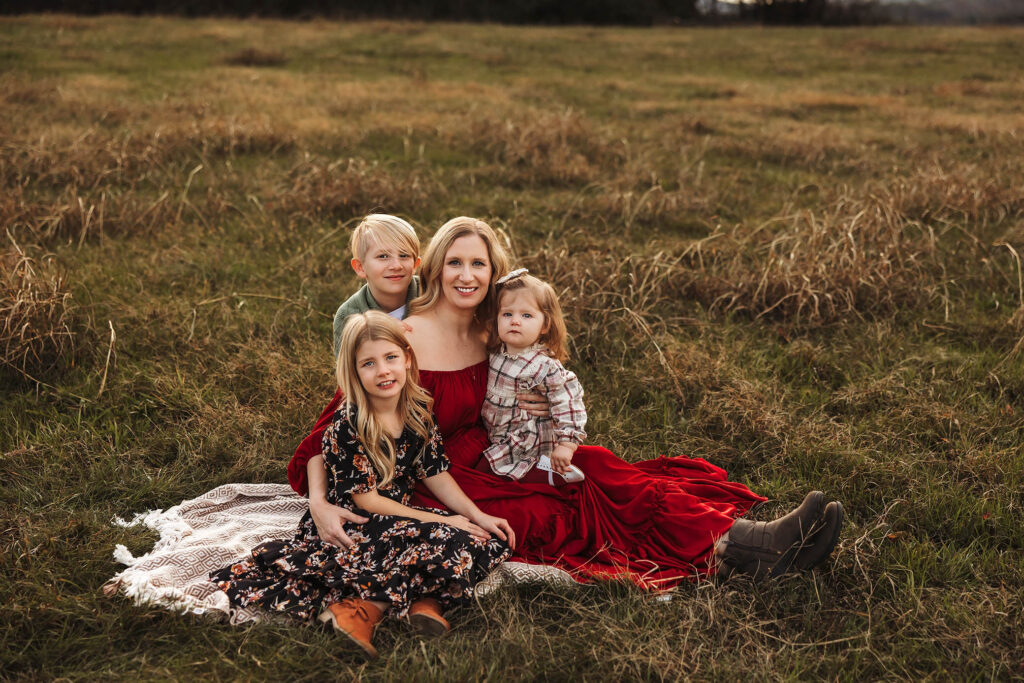 A woman in a red dress sits on a blanket in a grassy field with three smiling children, posing for a family photo by a talented Scottsdale maternity photographer.