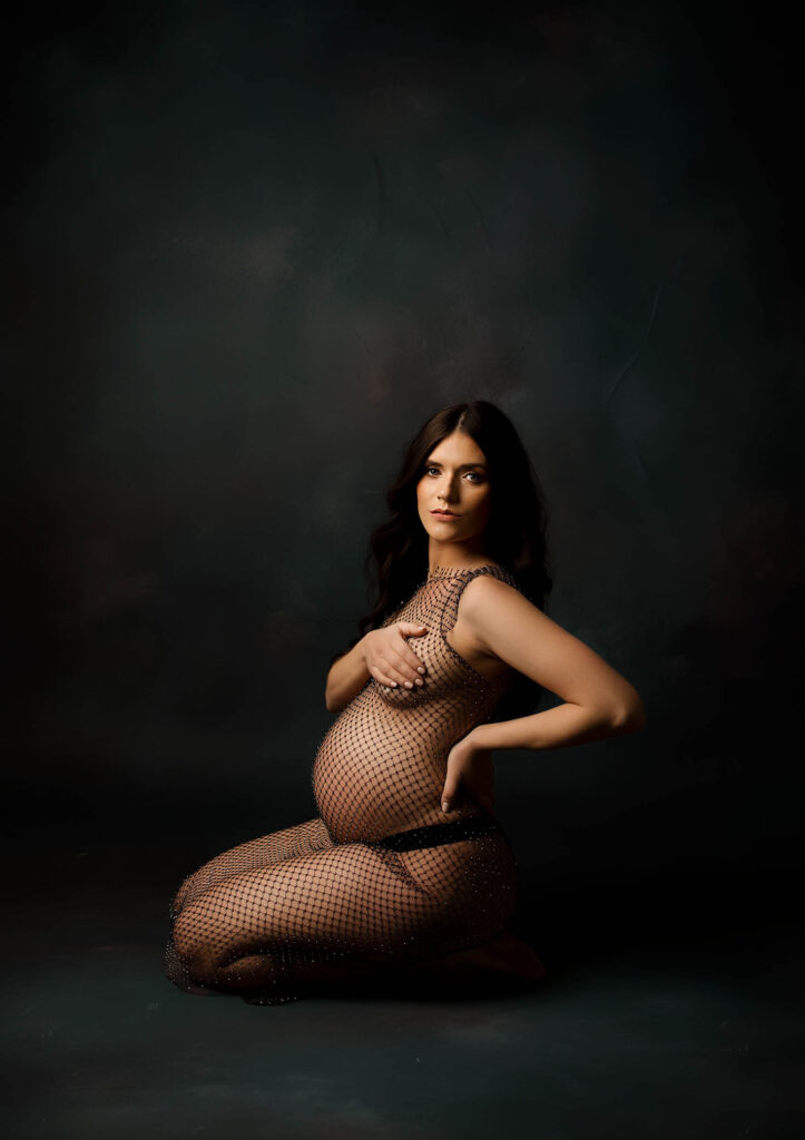 A pregnant woman kneels on the floor against a dark, textured background, wearing a sheer, netted dress and covering her chest with one arm while resting her other hand on her hip—captured by a skilled Phoenix maternity photographer.