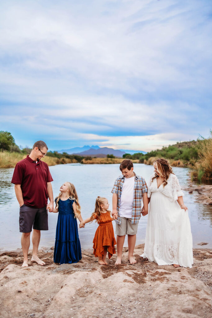 A family of five stands barefoot on a sandy riverbank, holding hands. Captured by a Phoenix maternity photographer, the father wears a maroon shirt and shorts, the mother a white dress, with their children in colorful outfits.