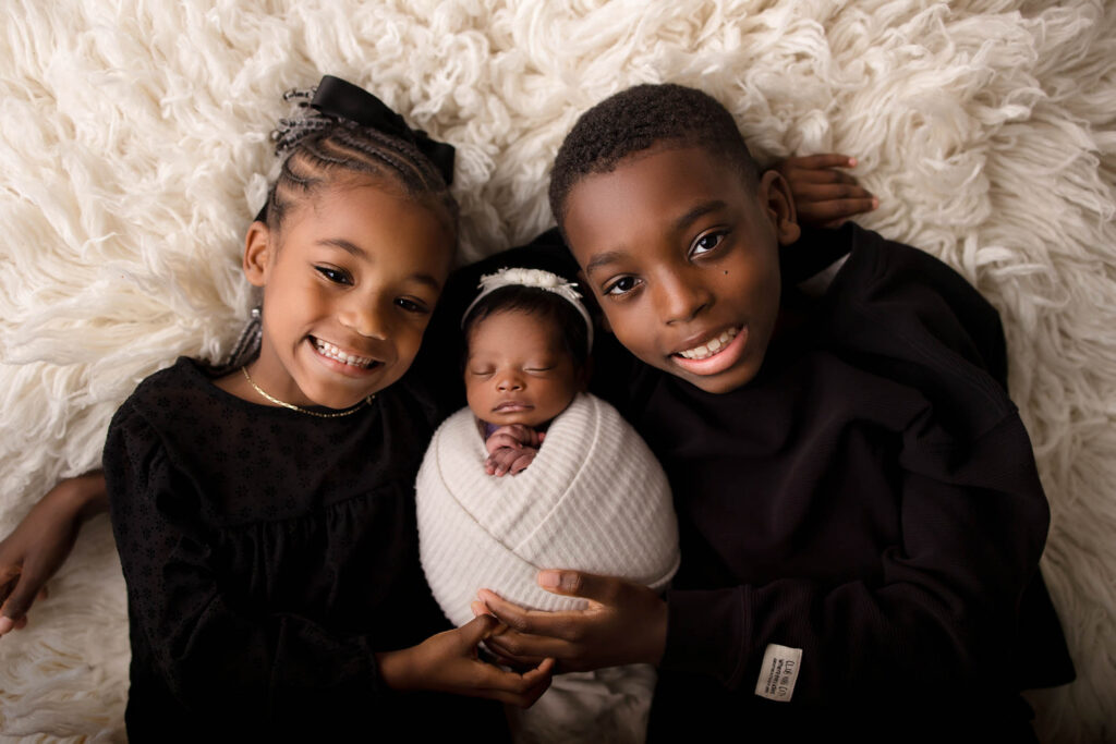 Two smiling children, a girl and a boy, lay on a fluffy white rug, holding a swaddled newborn baby between them—a heartwarming moment beautifully captured by a family photographer Phoenix AZ.