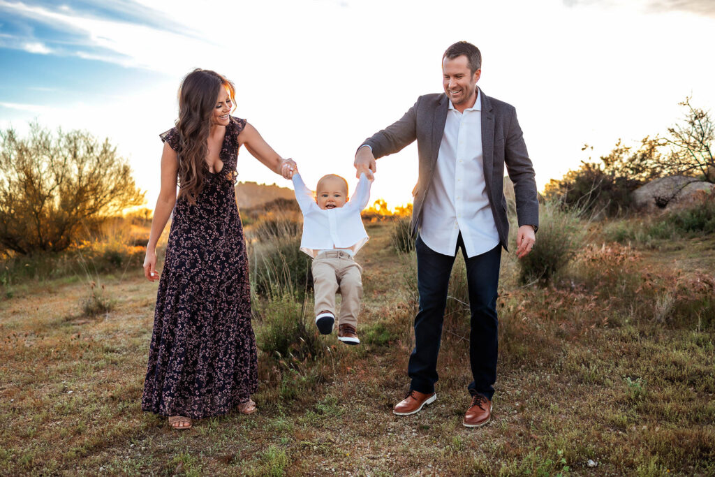 A smiling couple holds their young child by the hands, swinging him playfully in the air as a phoenix maternity photographer captures this joyful moment outdoors in a grassy field at sunset.
