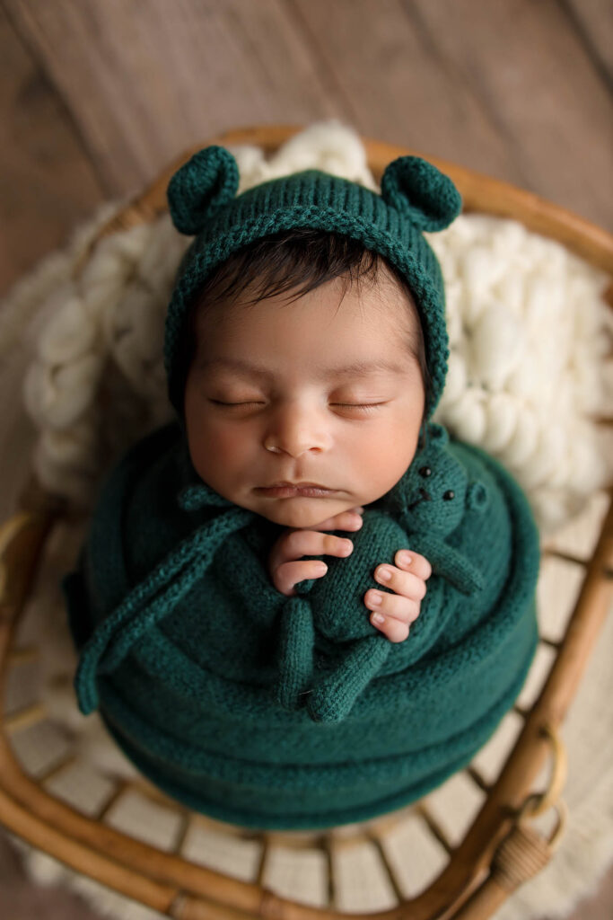 A newborn baby with dark hair sleeps peacefully, wrapped in a deep green blanket and matching knit hat with bear ears, holding a small teddy bear—captured by a skilled Phoenix newborn photographer—lying on a wooden chair with a soft cream blanket.