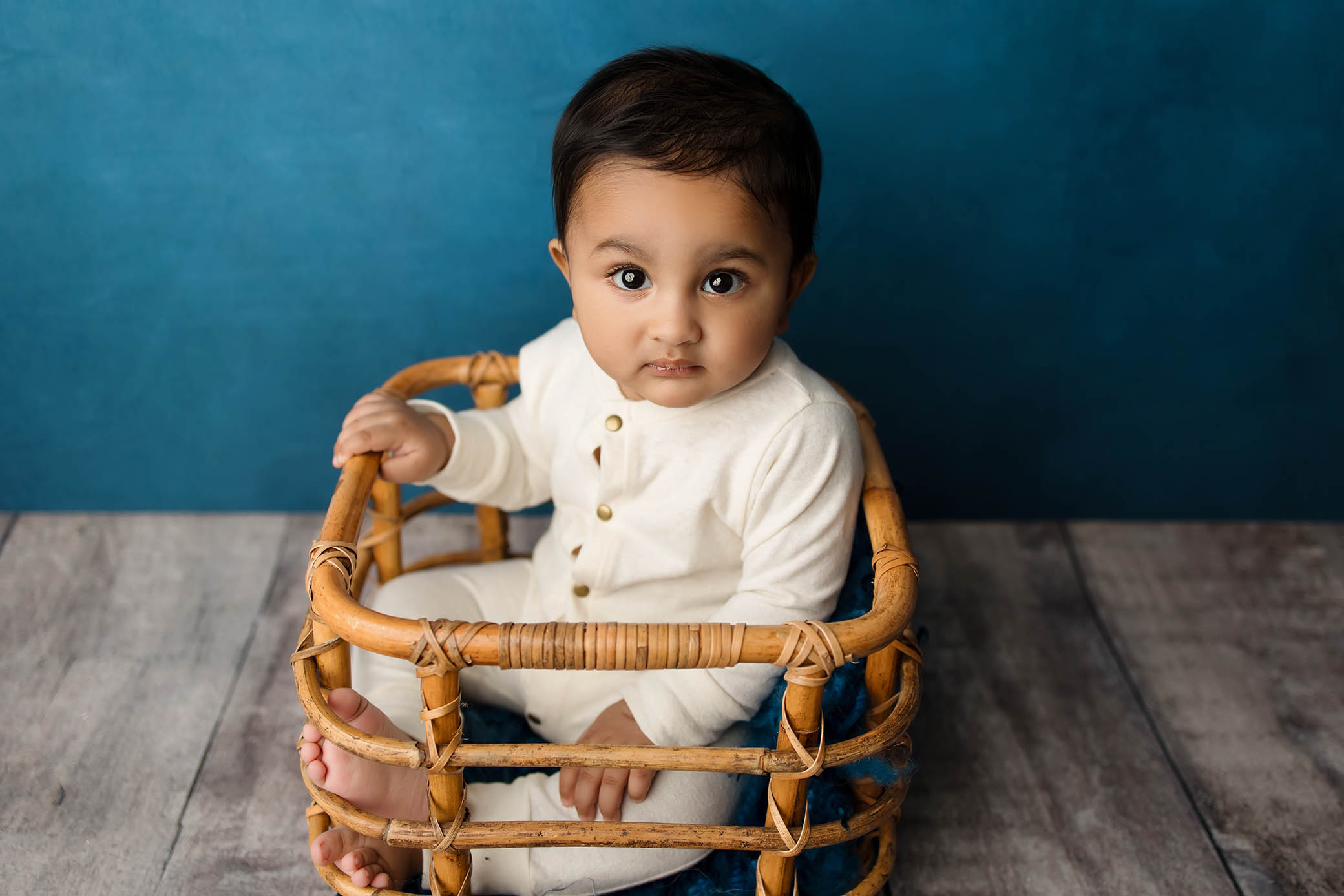 Baby in white outfit sits in woven basket on wooden floor, blue background, looking up.