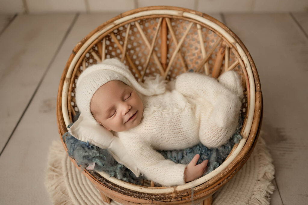 A newborn baby dressed in a white knit outfit and hat smiles while sleeping in a round wicker basket, captured by a Phoenix maternity photographer, lying on a small pillow and blue blanket on a wooden floor.
