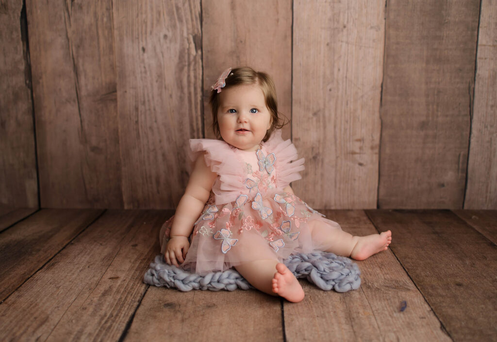A baby with light brown hair and blue eyes sits on a gray knitted blanket, wearing a pink dress with butterfly appliqués and tulle ruffles, in front of a wooden backdrop—perfect for a family photographer Phoenix AZ session.