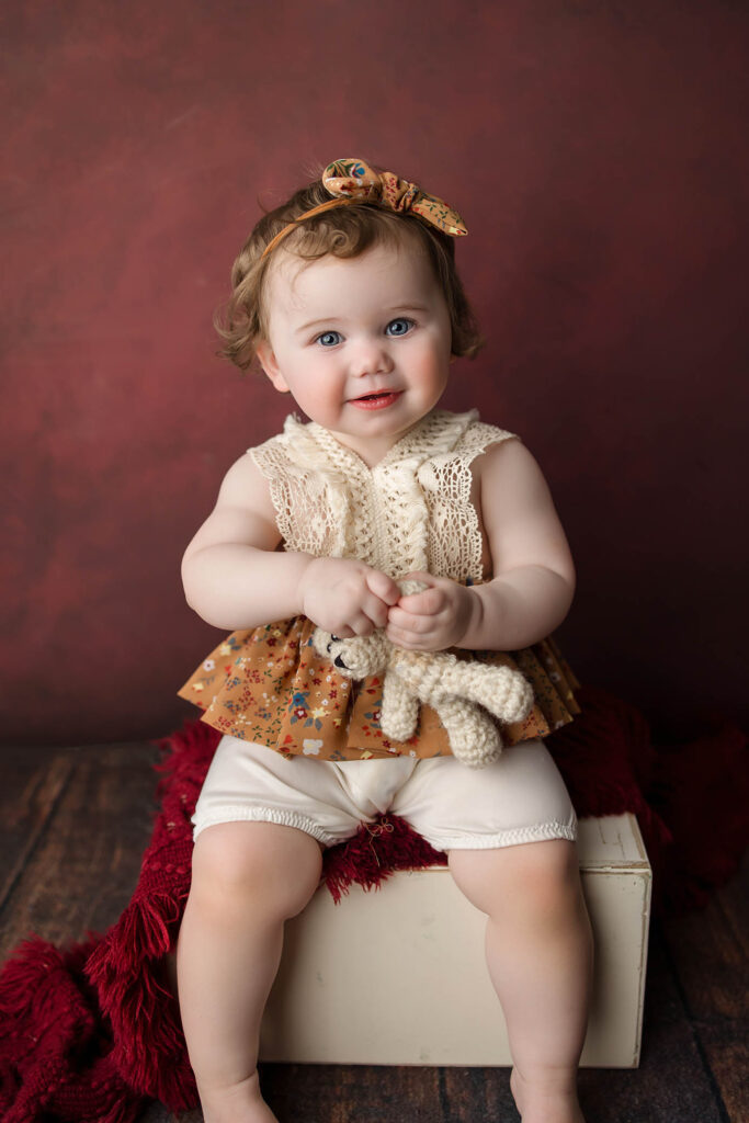 A smiling toddler with blue eyes sits on a white box against a maroon background, wearing a floral headband and lace top, holding a small knitted toy—a heartwarming moment captured by a family photographer Phoenix AZ. A red textured blanket drapes over the box.