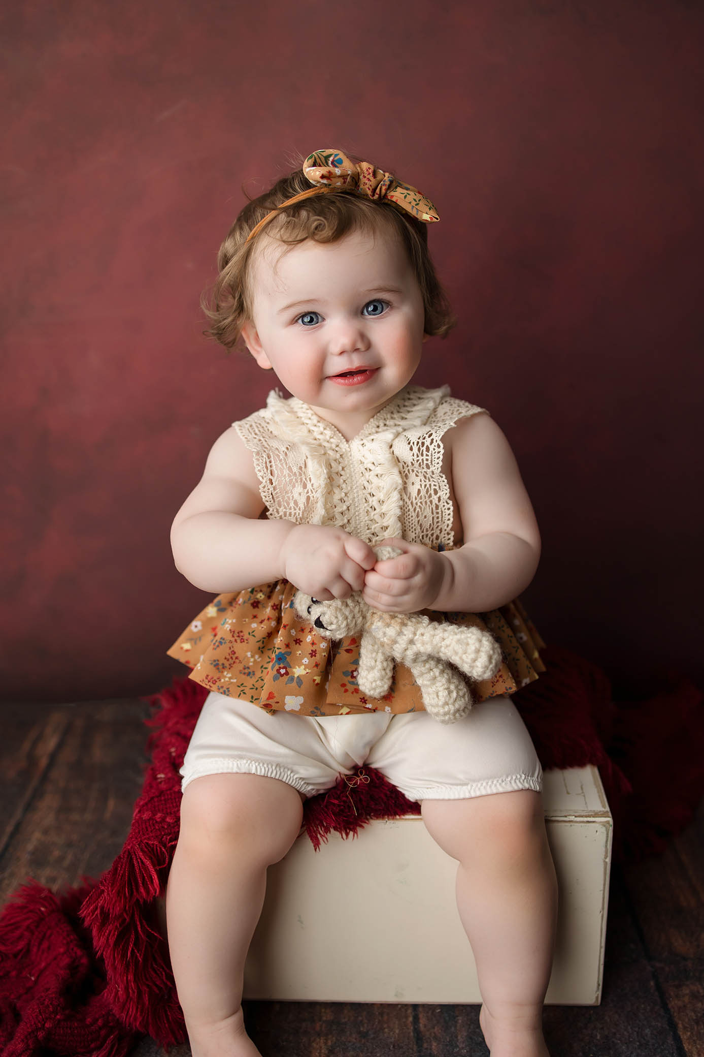 A smiling toddler with blue eyes sits on a white box against a maroon background, wearing a floral headband and lace top, holding a small knitted toy—a heartwarming moment captured by a family photographer Phoenix AZ. A red textured blanket drapes over the box.