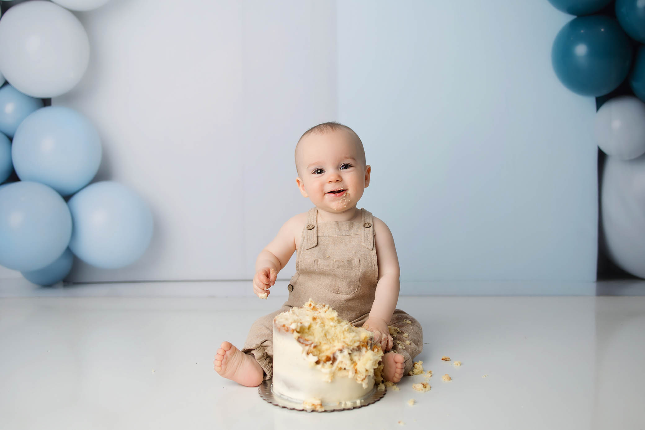Smiling baby in beige overalls with smashed cake, surrounded by light blue and white balloons.